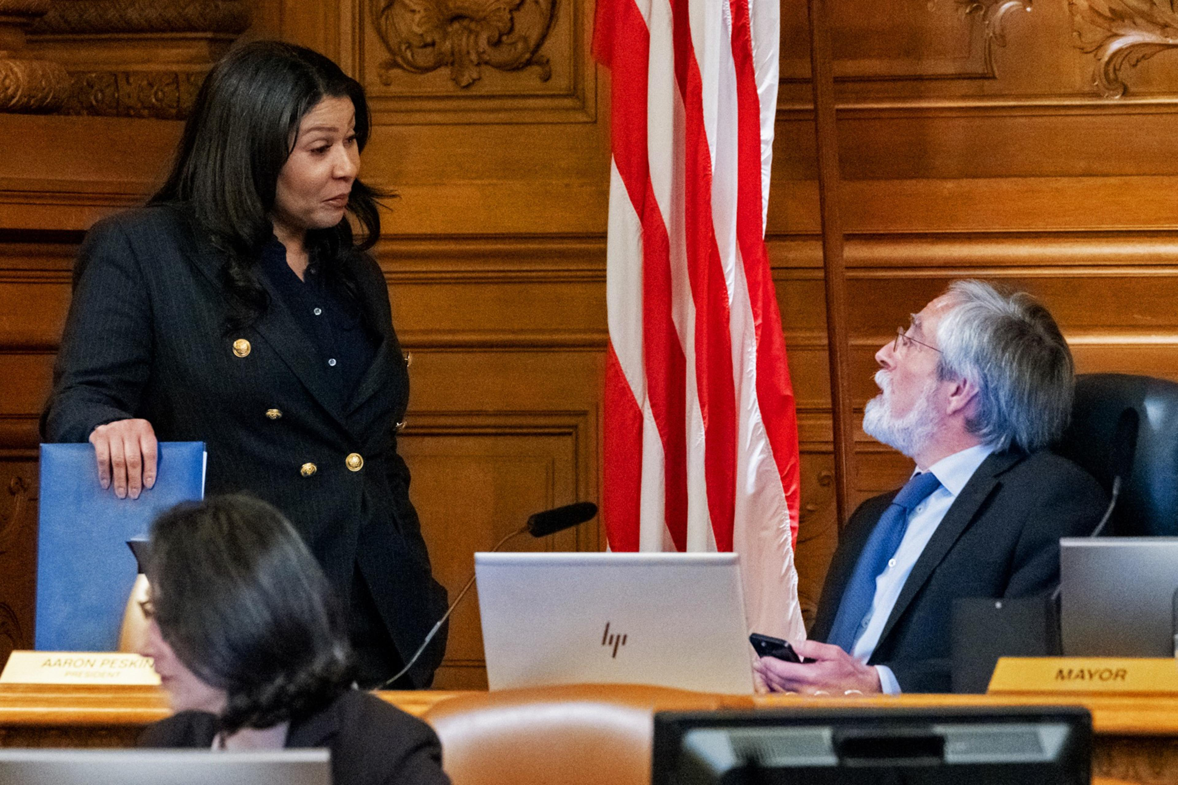 A woman converses with a seated man; both are in a wood-paneled room with a US flag.