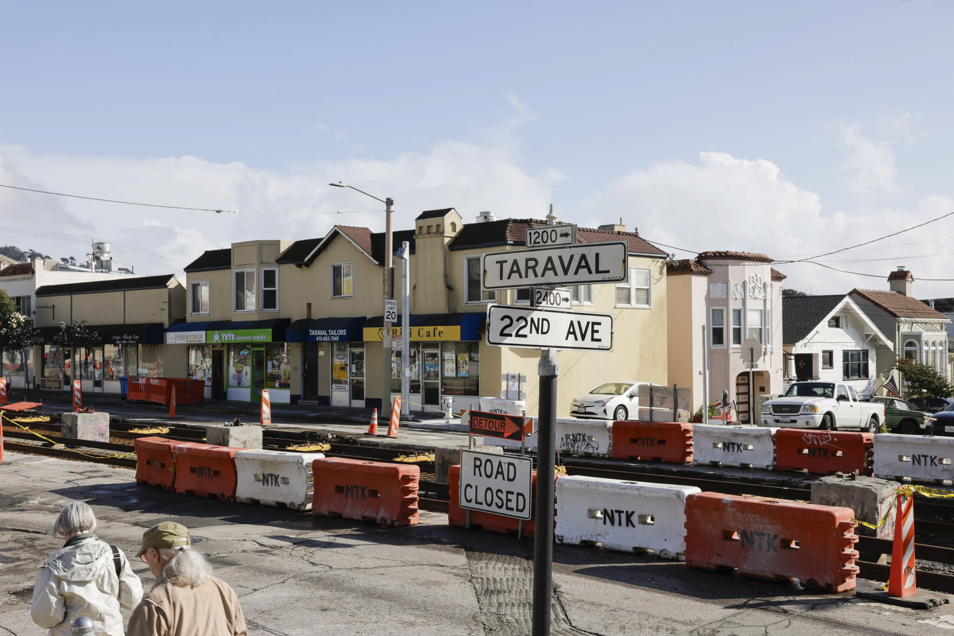 A street corner with road closure barriers, detour sign, and two people observing the scene. Shops line the street in the background.