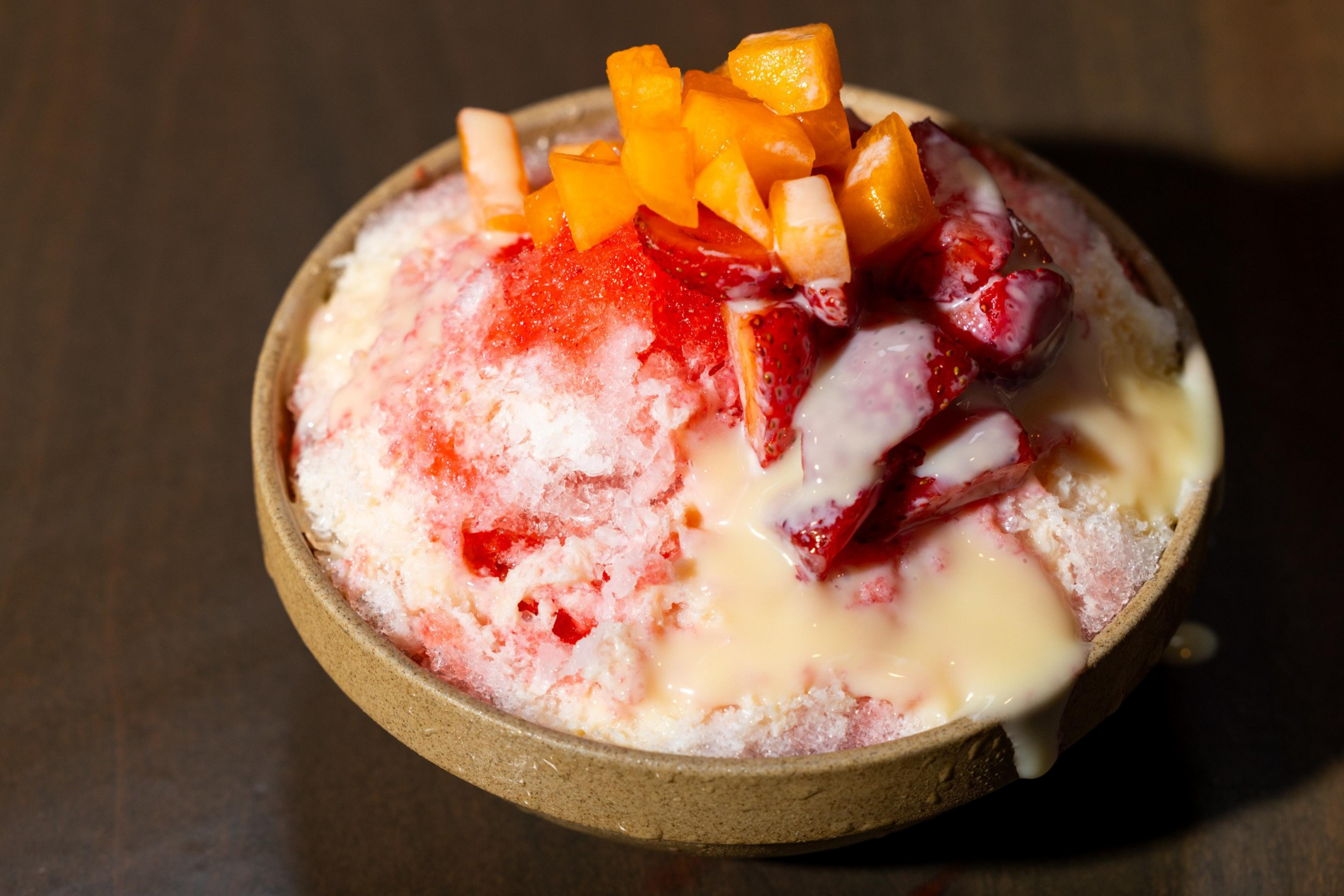 A bowl of shaved ice topped with red syrup, custard-like cream, sliced strawberries, and small orange fruit cubes.