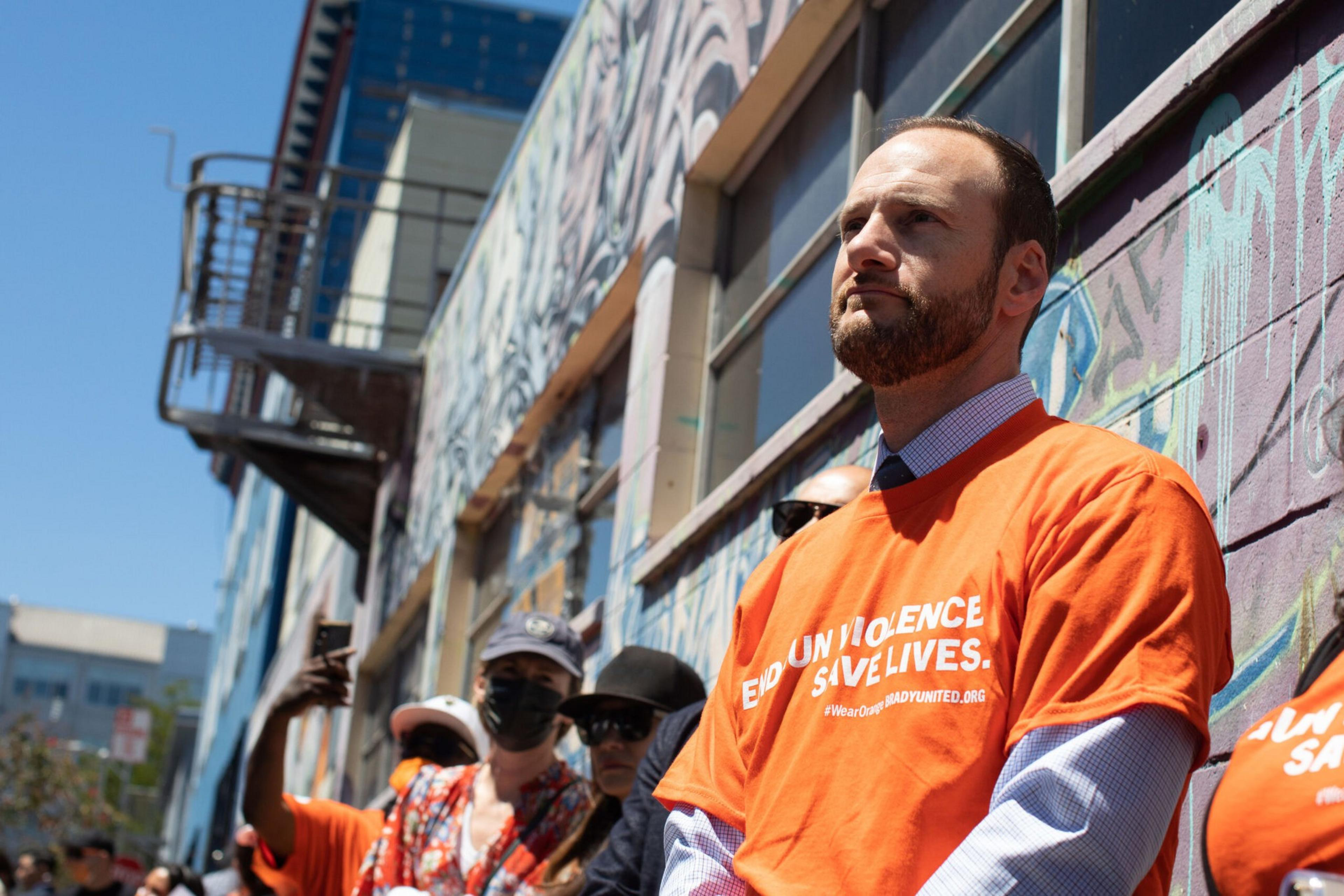 A man in an orange shirt that says "END GUN VIOLENCE SAVE LIVES" stands solemnly among others against a graffiti-covered wall on a sunny day.