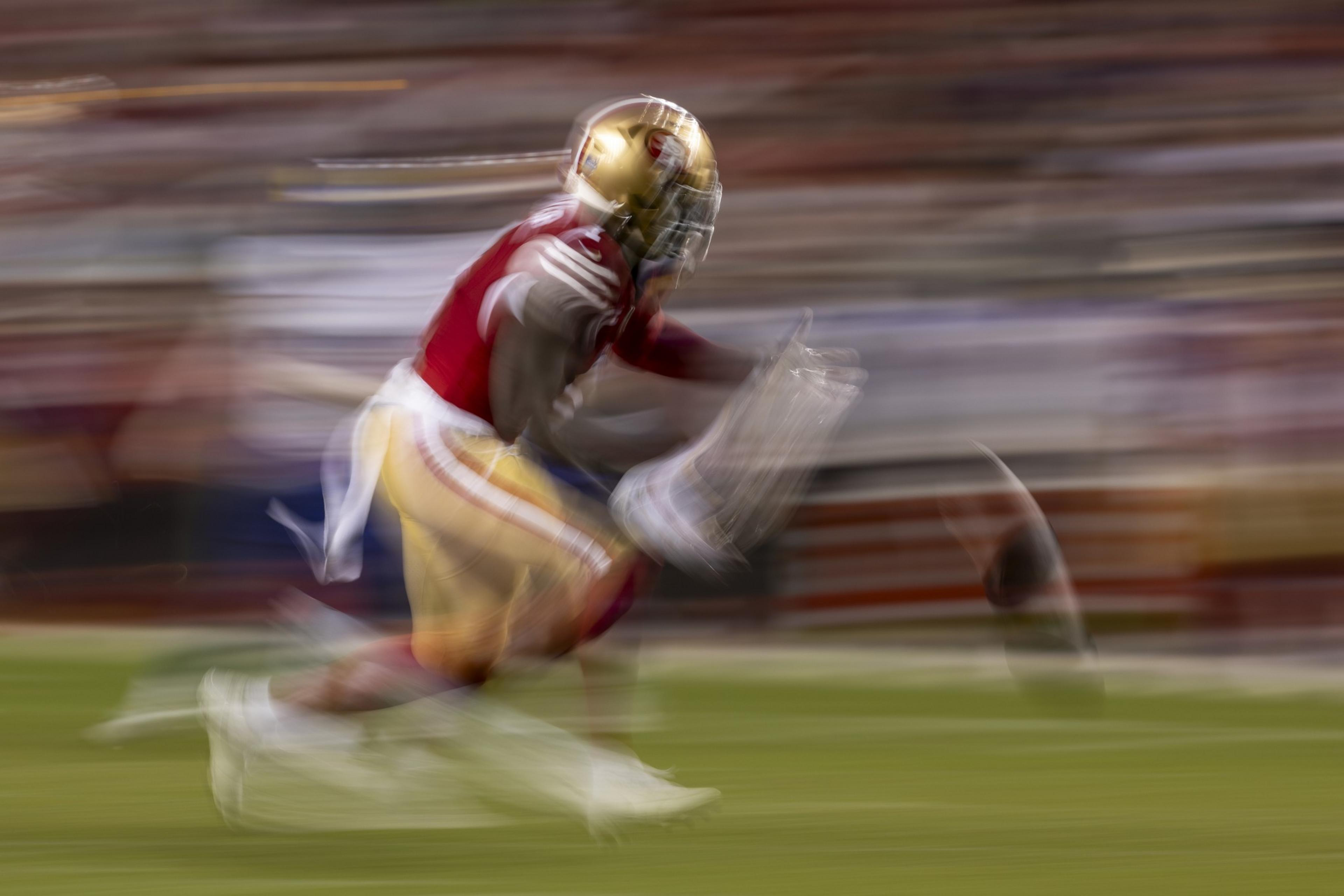 A football player in red and gold, blurred in motion, is running quickly on a field. The background is streaked, indicating speed.