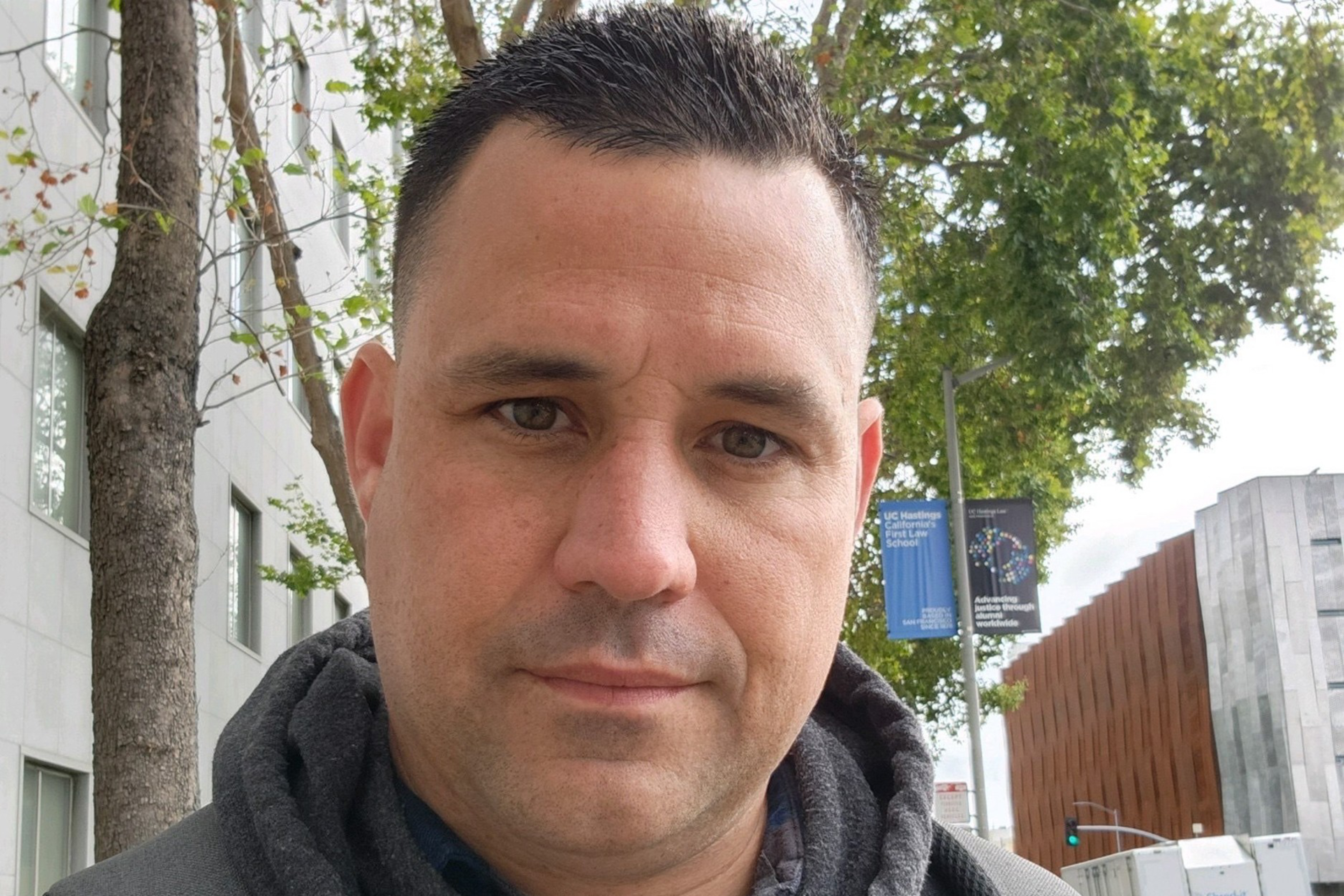 A man with short dark hair and brown eyes looks directly at the camera, wearing a dark hoodie, standing outdoors near trees and buildings.
