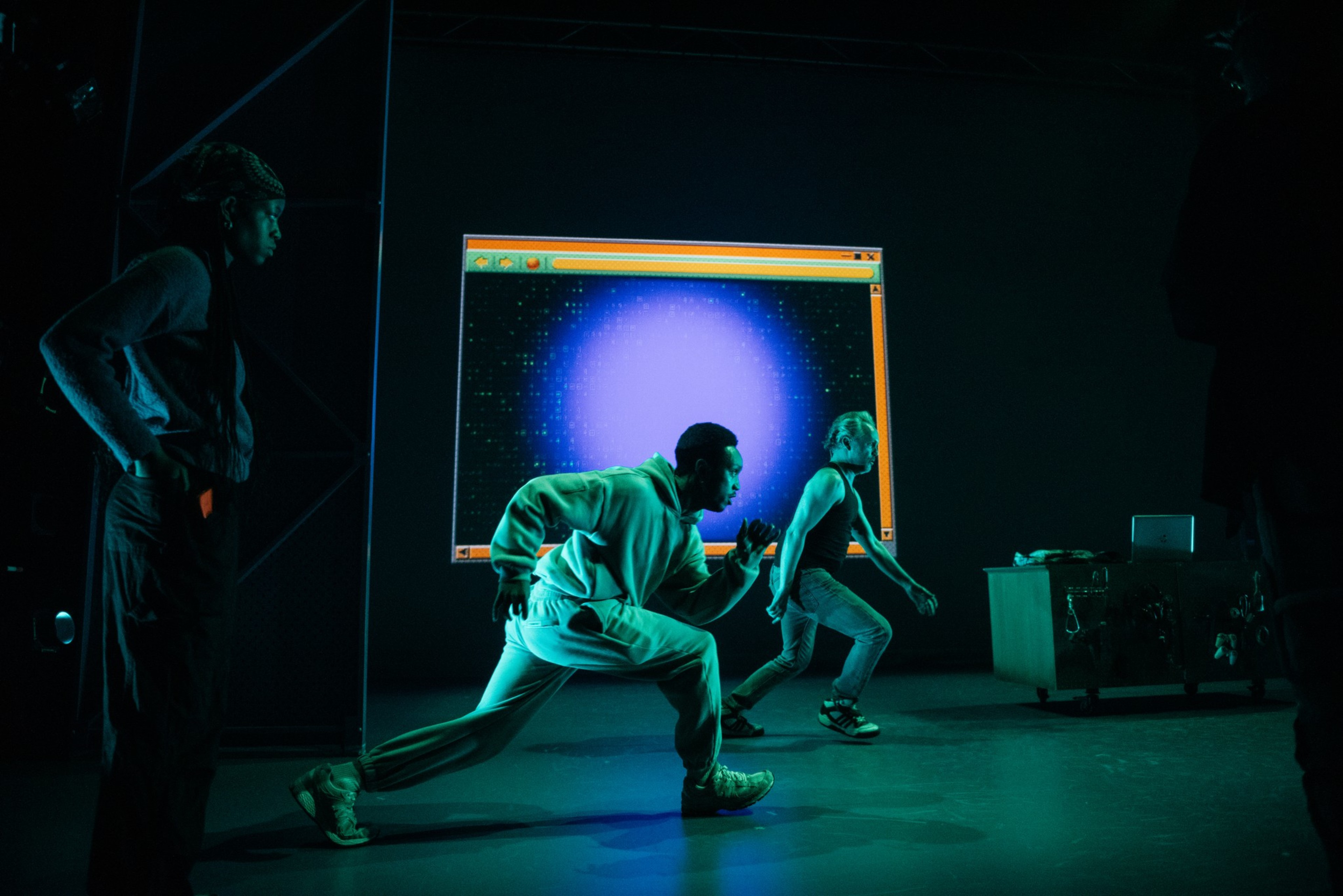 Two people in dynamic poses appear in front of a glowing screen resembling a computer window, with a blue and purple light effect, in a dimly lit setting.