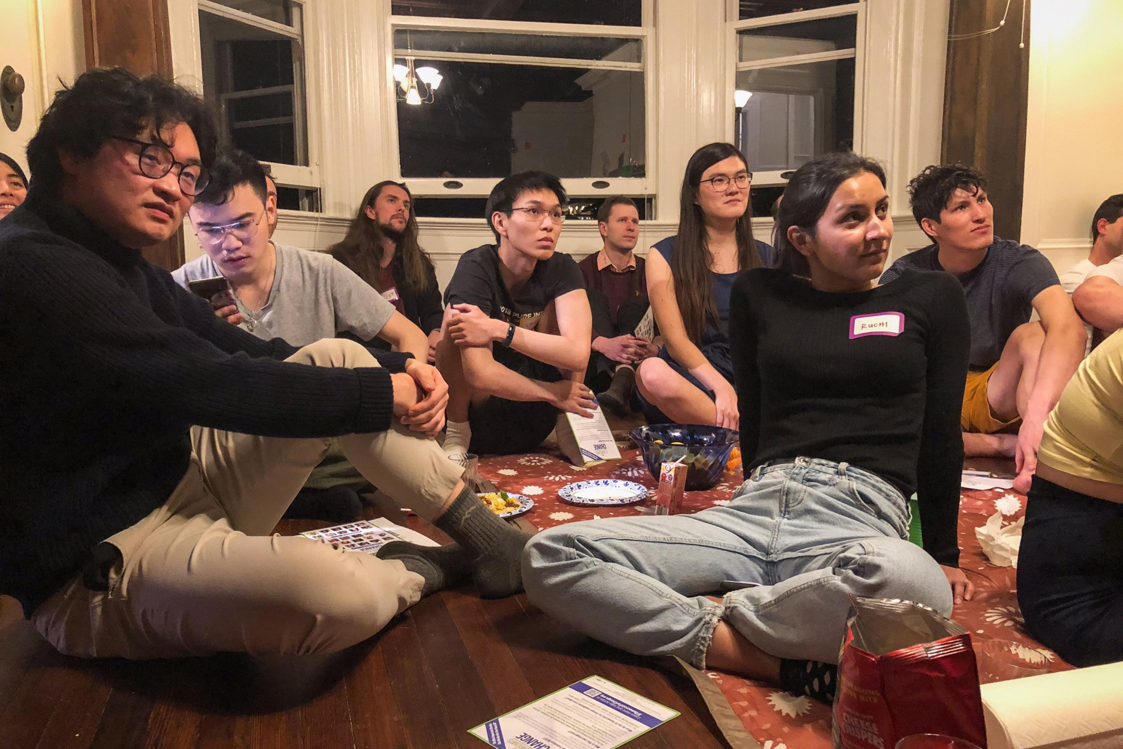 A group of young adults sit on the floor in a Victorian-style apartment.