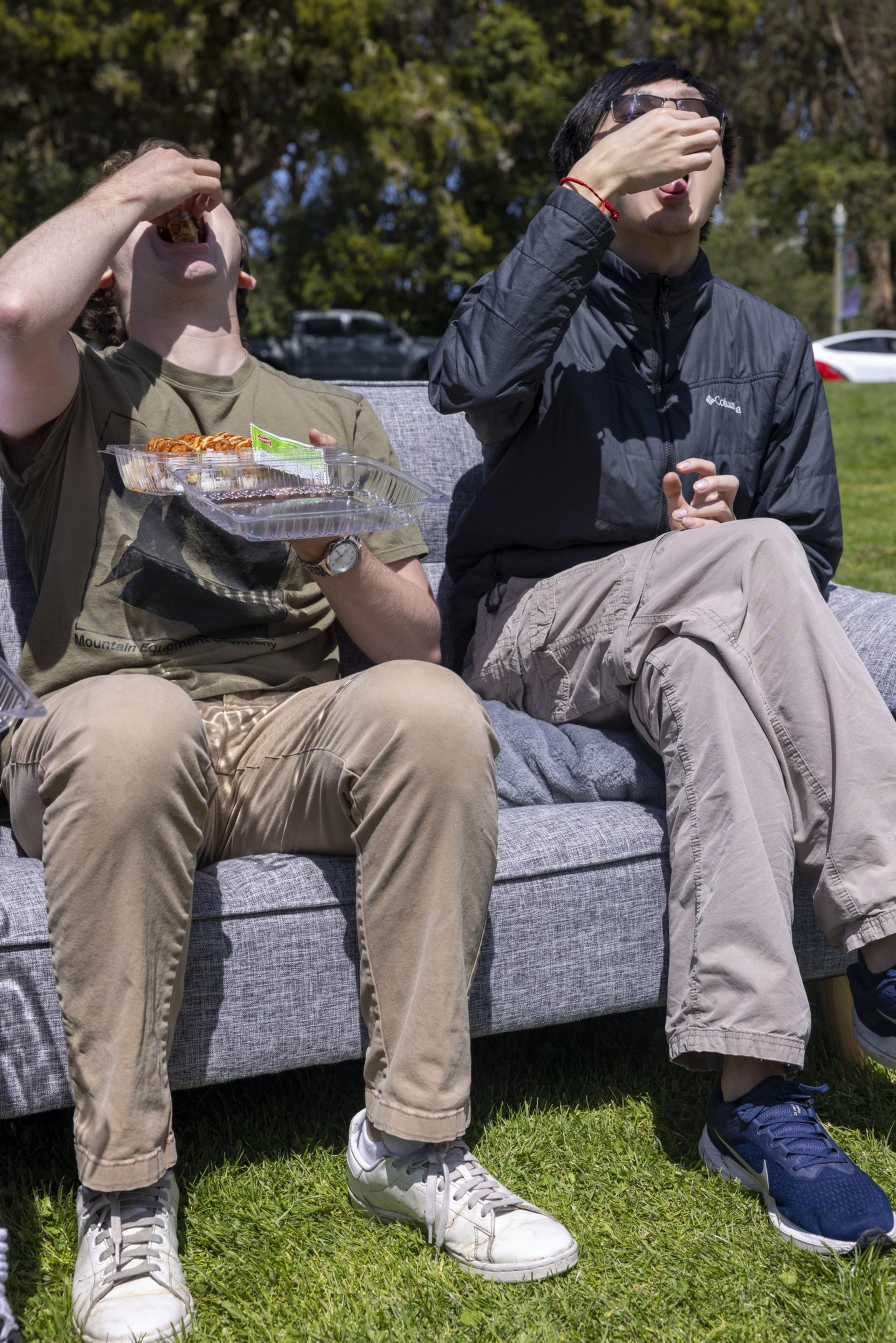Two people sit on a gray outdoor sofa, eating food from a plastic tray on a sunny day. The grassy area and trees are visible in the background.
