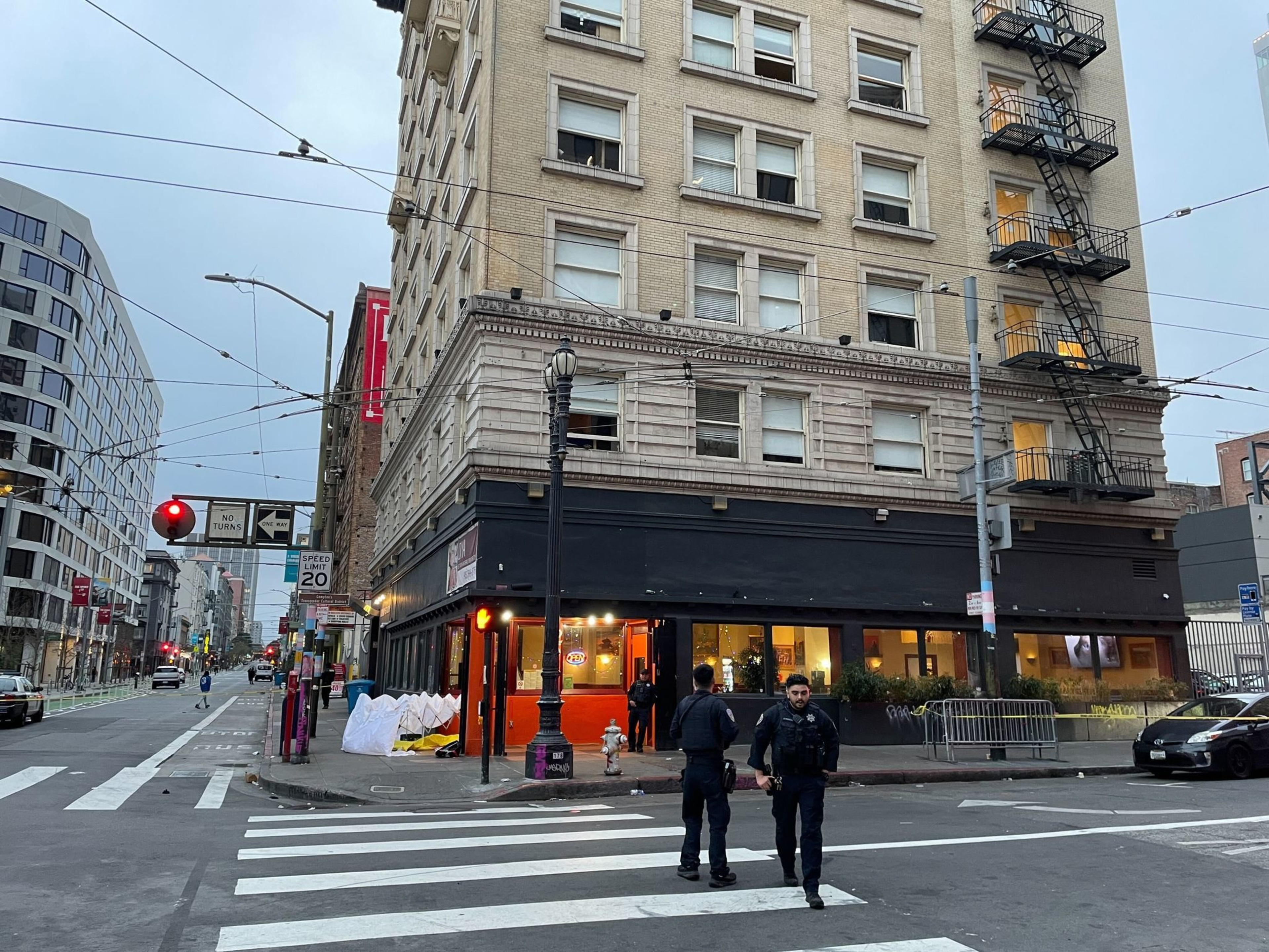 Two police officers are walking across a city intersection. They're near a multi-story building with a fire escape, and traffic signs are visible.