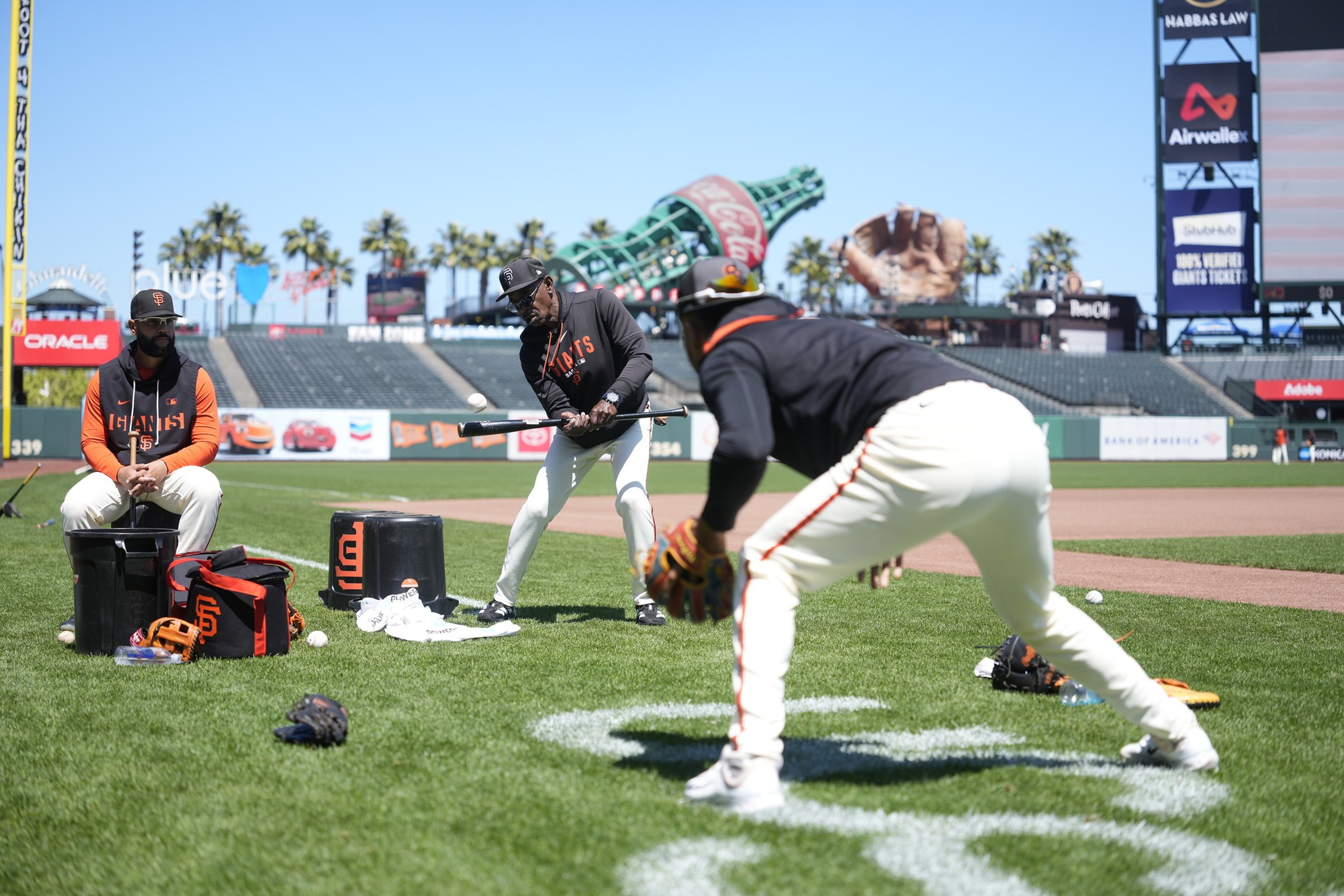 Three baseball players in Giants uniforms practice on a field, one batting, one fielding, and one sitting on a bucket with gear around.
