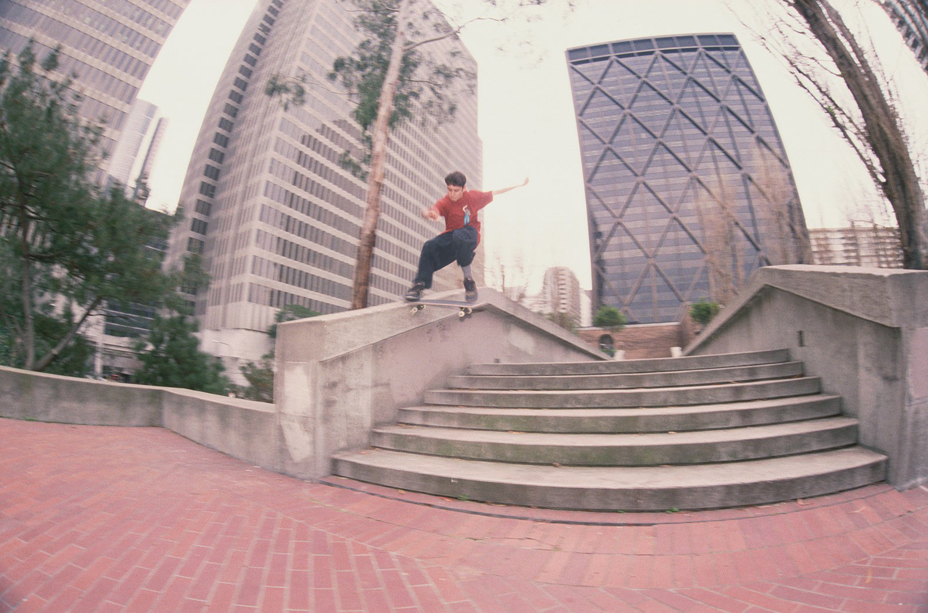 A skateboarder in a red shirt grinds on a concrete ledge above steps in an urban plaza with high-rise buildings and trees in the background.