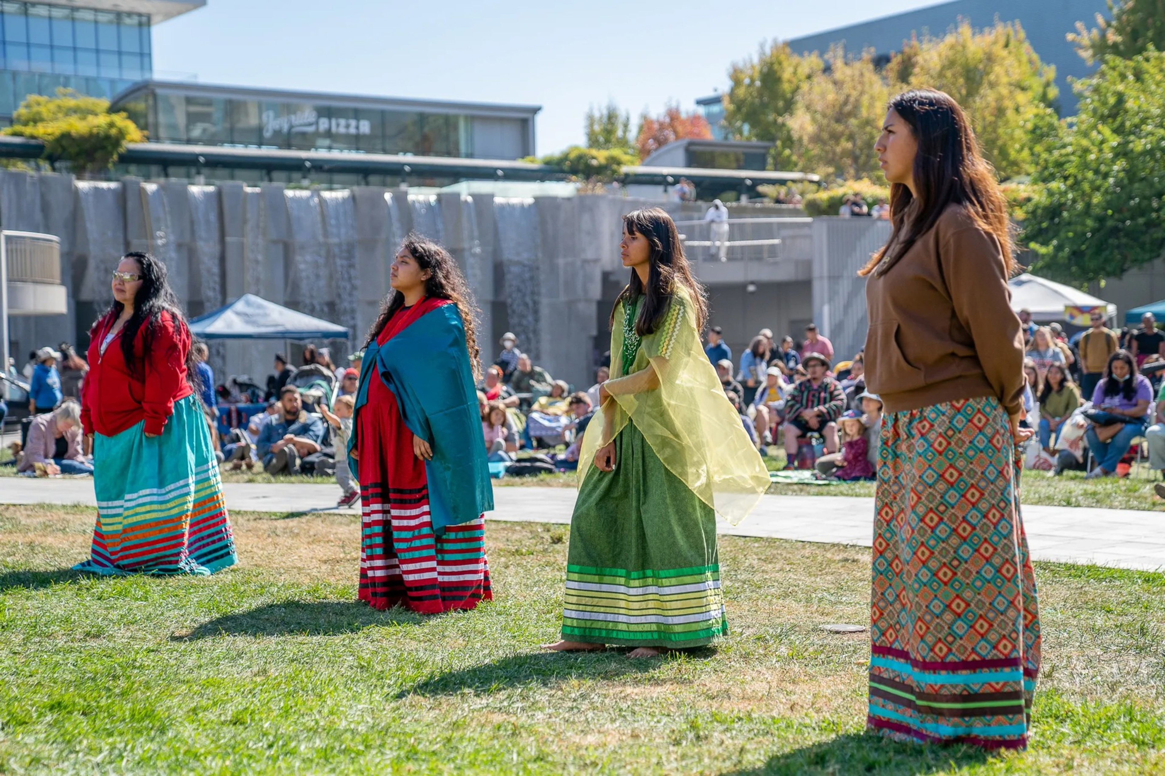 Four women stand spaced apart on grass, wearing colorful traditional skirts and shawls, with a seated crowd and a waterfall feature in the background.