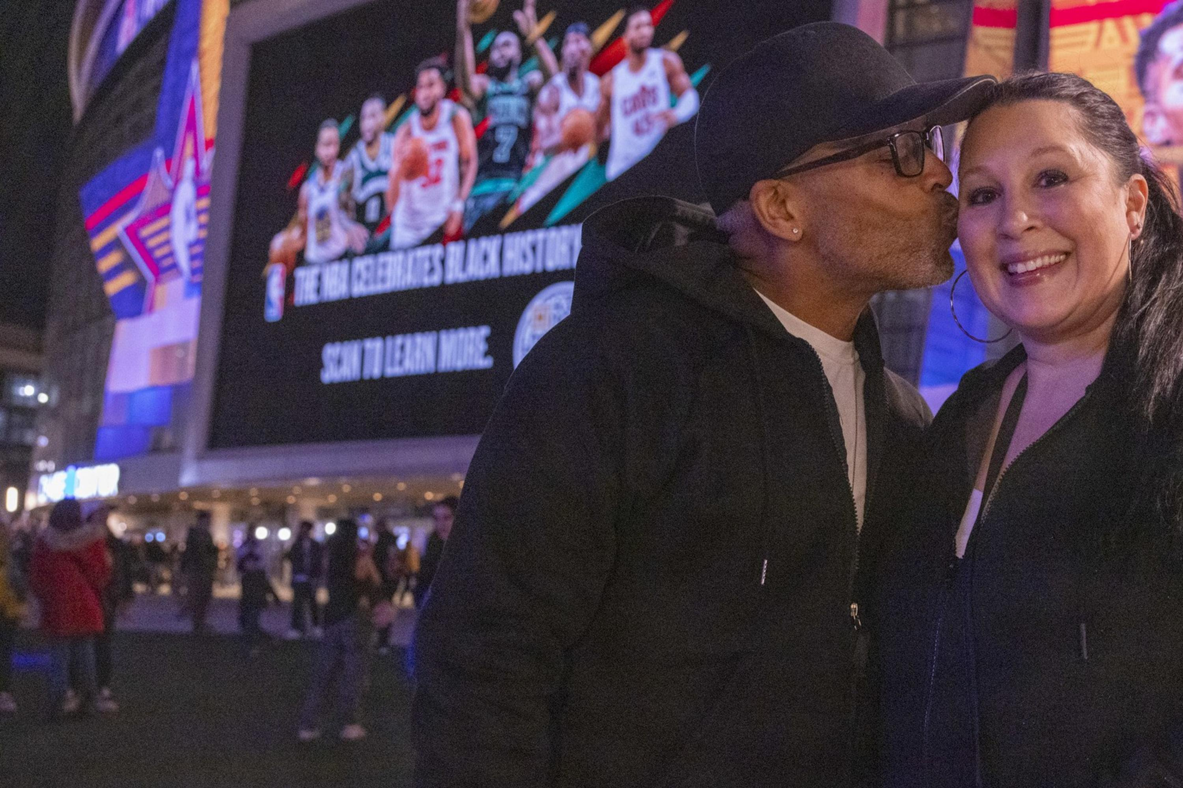 A man in a cap kisses a smiling woman on the cheek. They're outdoors near a large, colorful billboard displaying basketball players and text.