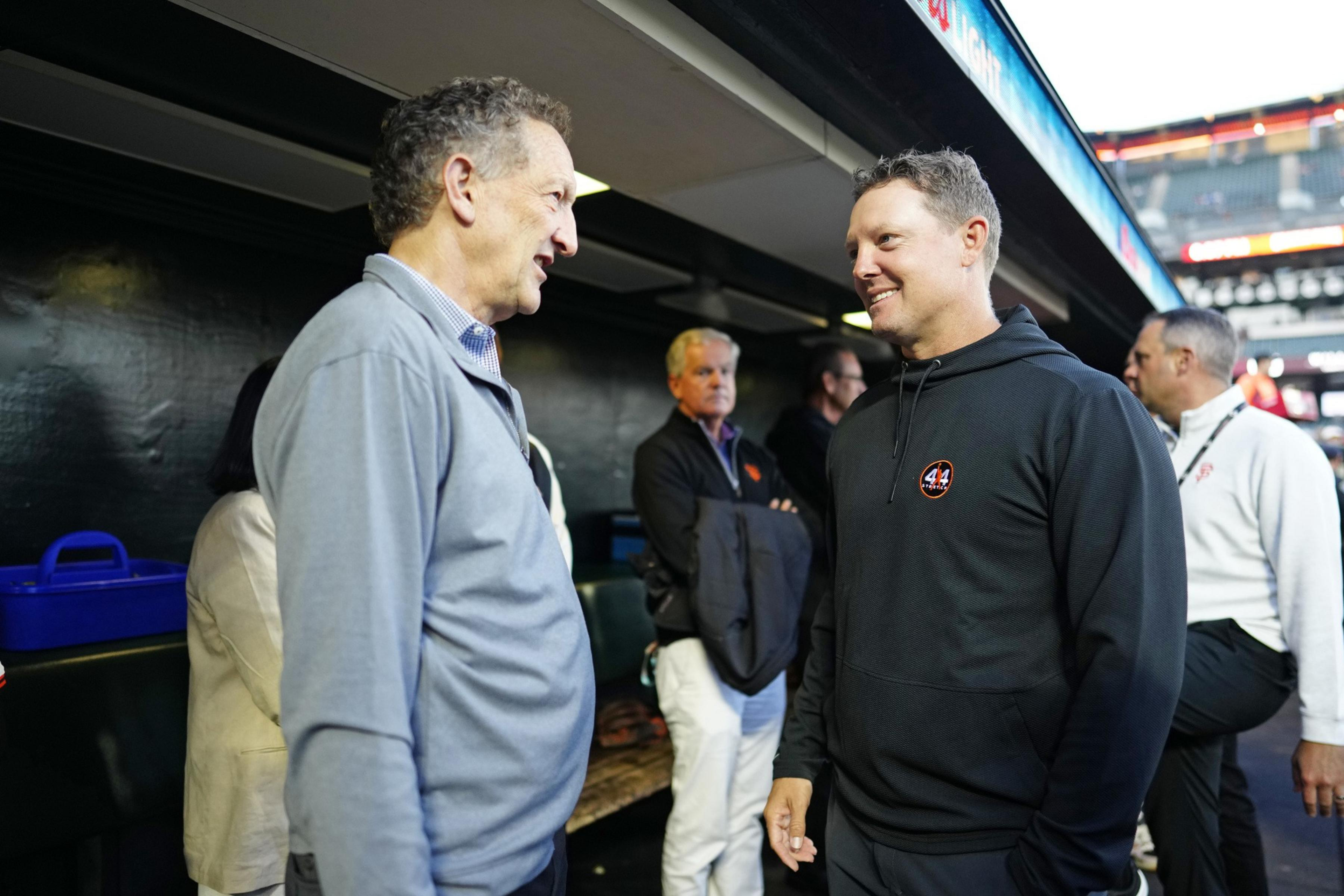 Two men, one in a gray jacket and the other in a black hoodie, are smiling and conversing in what appears to be a sports dugout with other people in the background.
