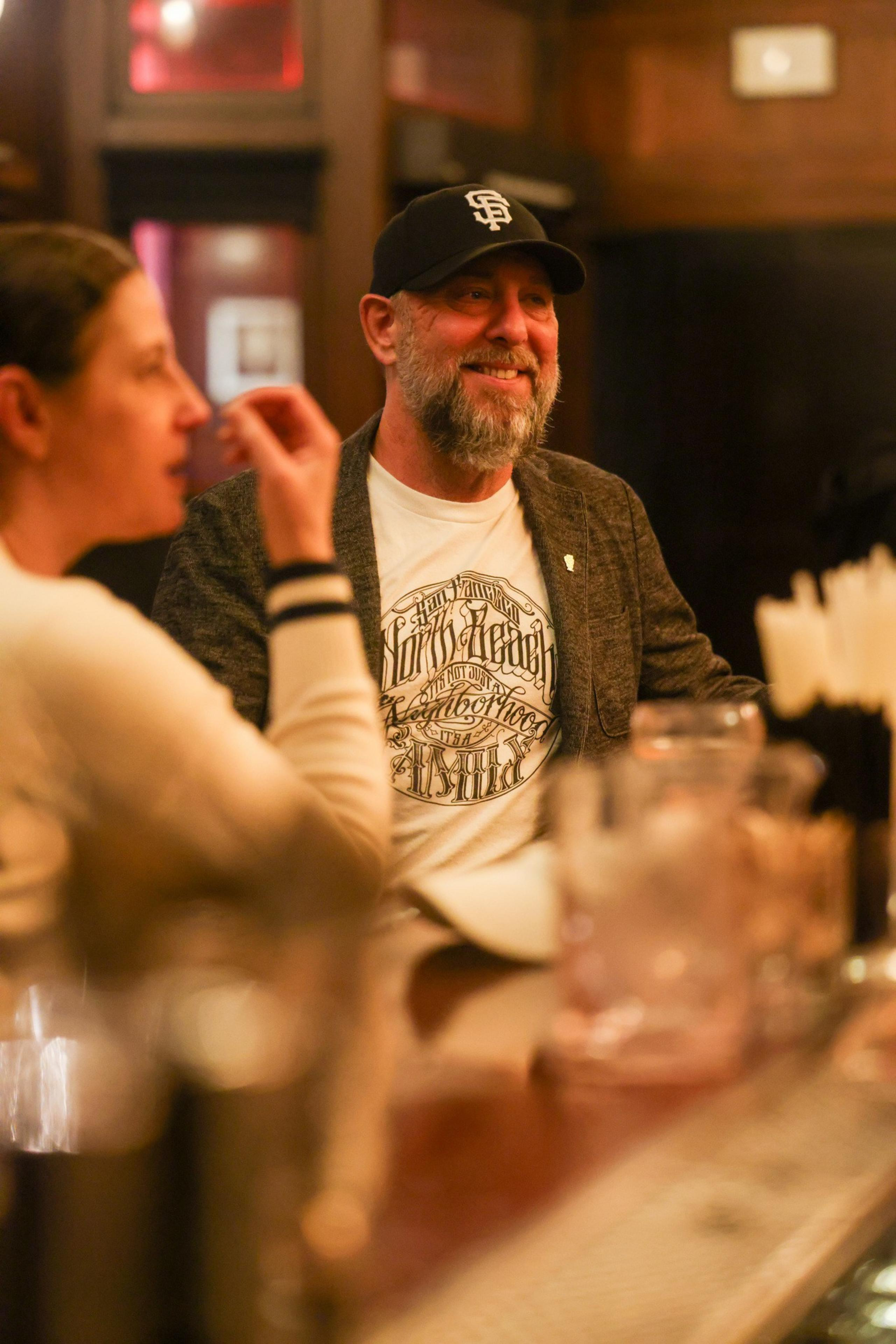 A bearded man in a cap and graphic tee smiles while sitting at a bar next to a woman in a white sweater, with blurred foreground details.
