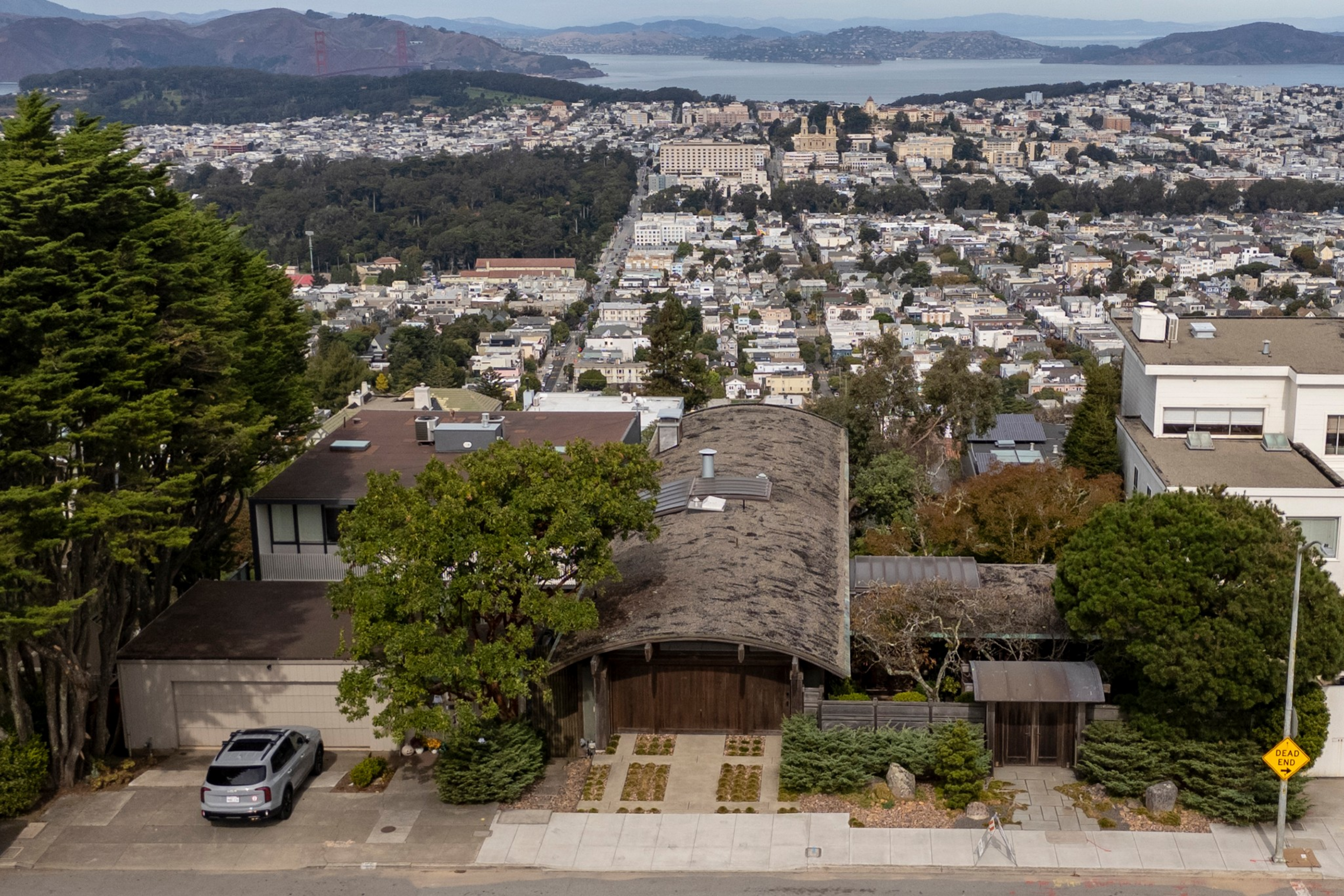 A residential area with a unique arched-roof house, surrounded by trees, overlooks a densely packed cityscape and distant bay with hills.