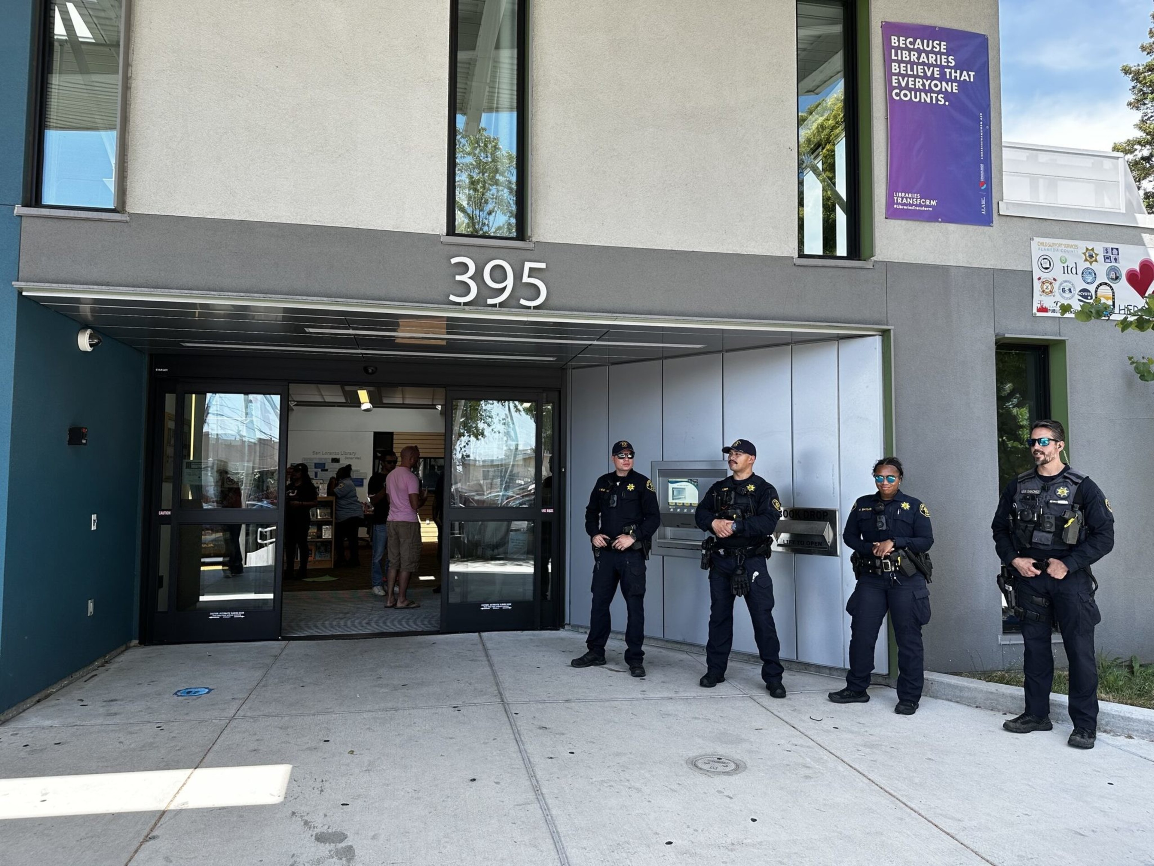 Four police officers in uniform stand outside the entrance of a building numbered 395, near a sign promoting library inclusivity.