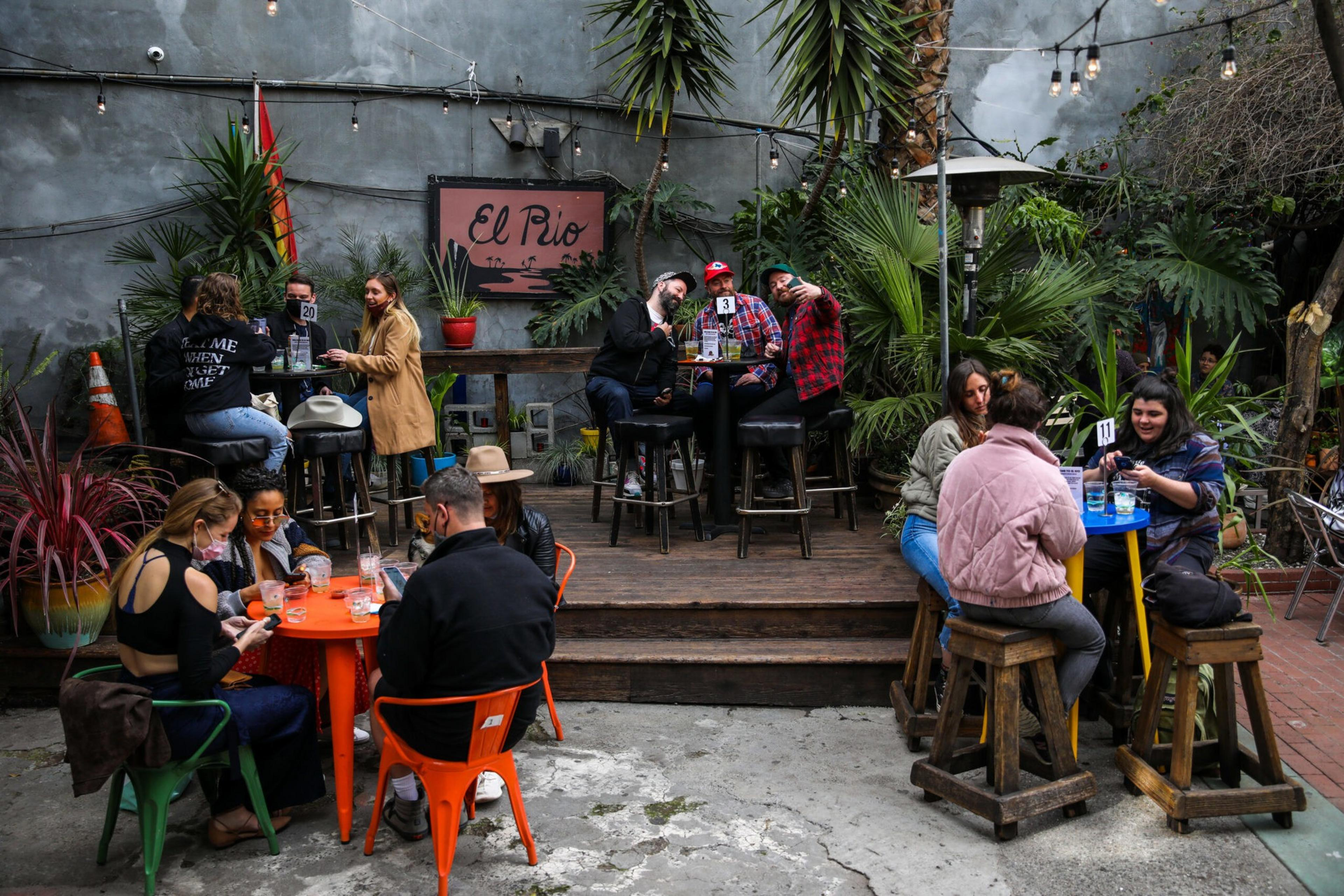 People are socializing at an outdoor venue called "El Rio" with plants, tables, and string lights decorating the area.