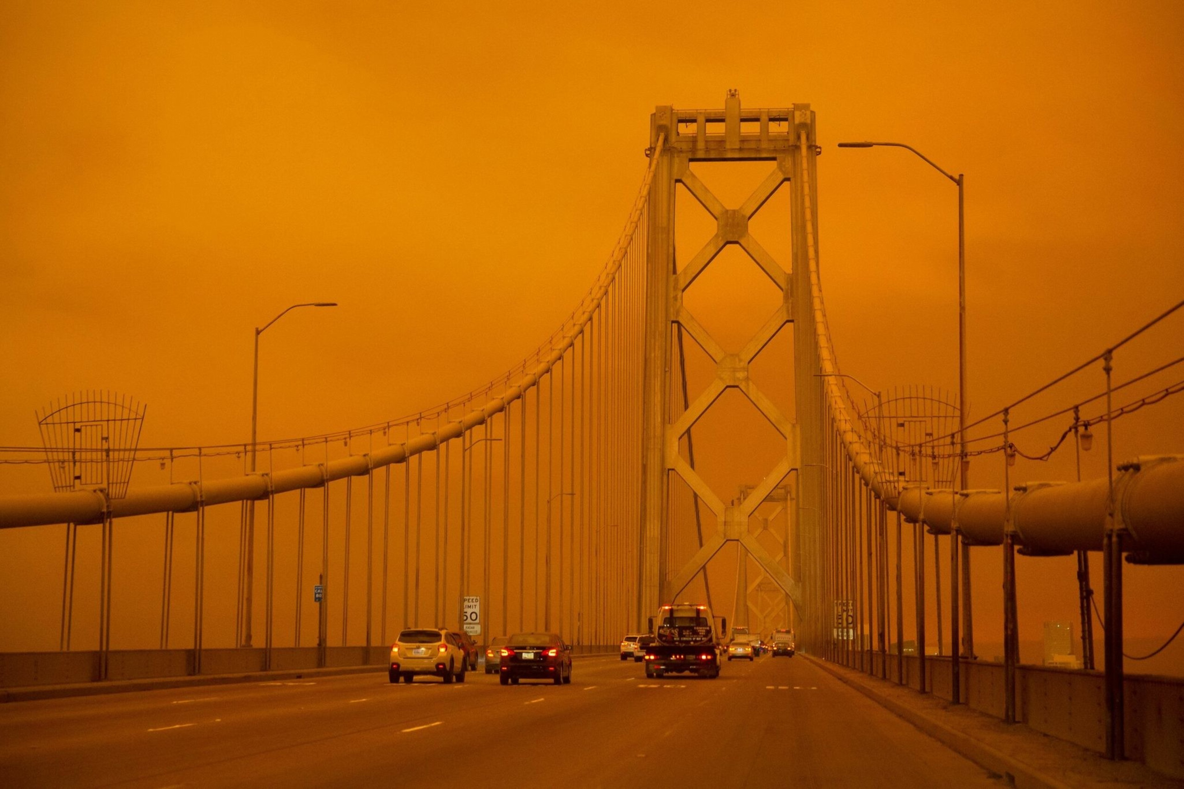 A suspension bridge with several vehicles driving across under an intense orange sky, likely caused by atmospheric conditions.