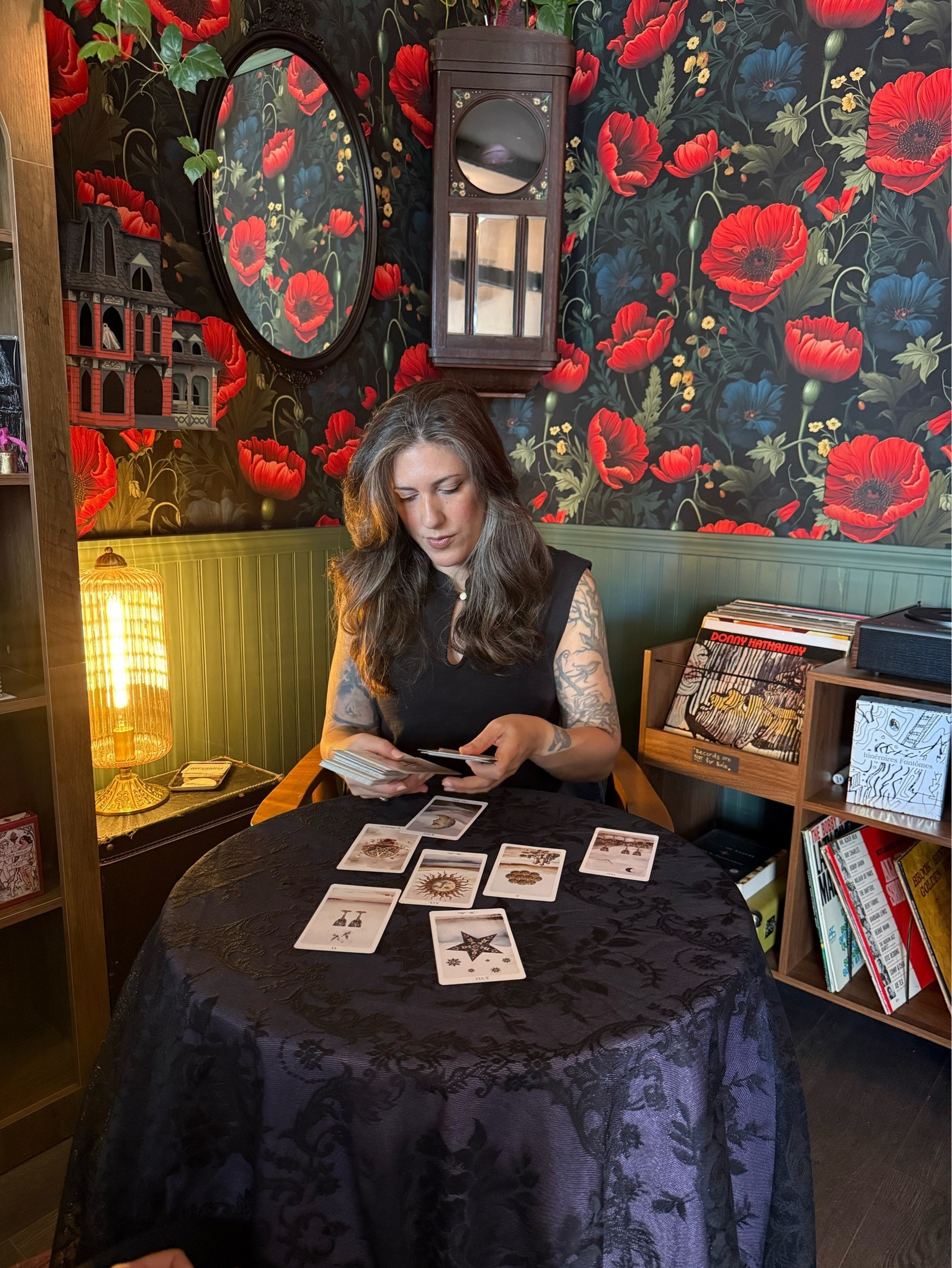 A woman with long hair and tattoos sits at a round table with tarot cards spread out, surrounded by floral wallpaper and vintage decor.
