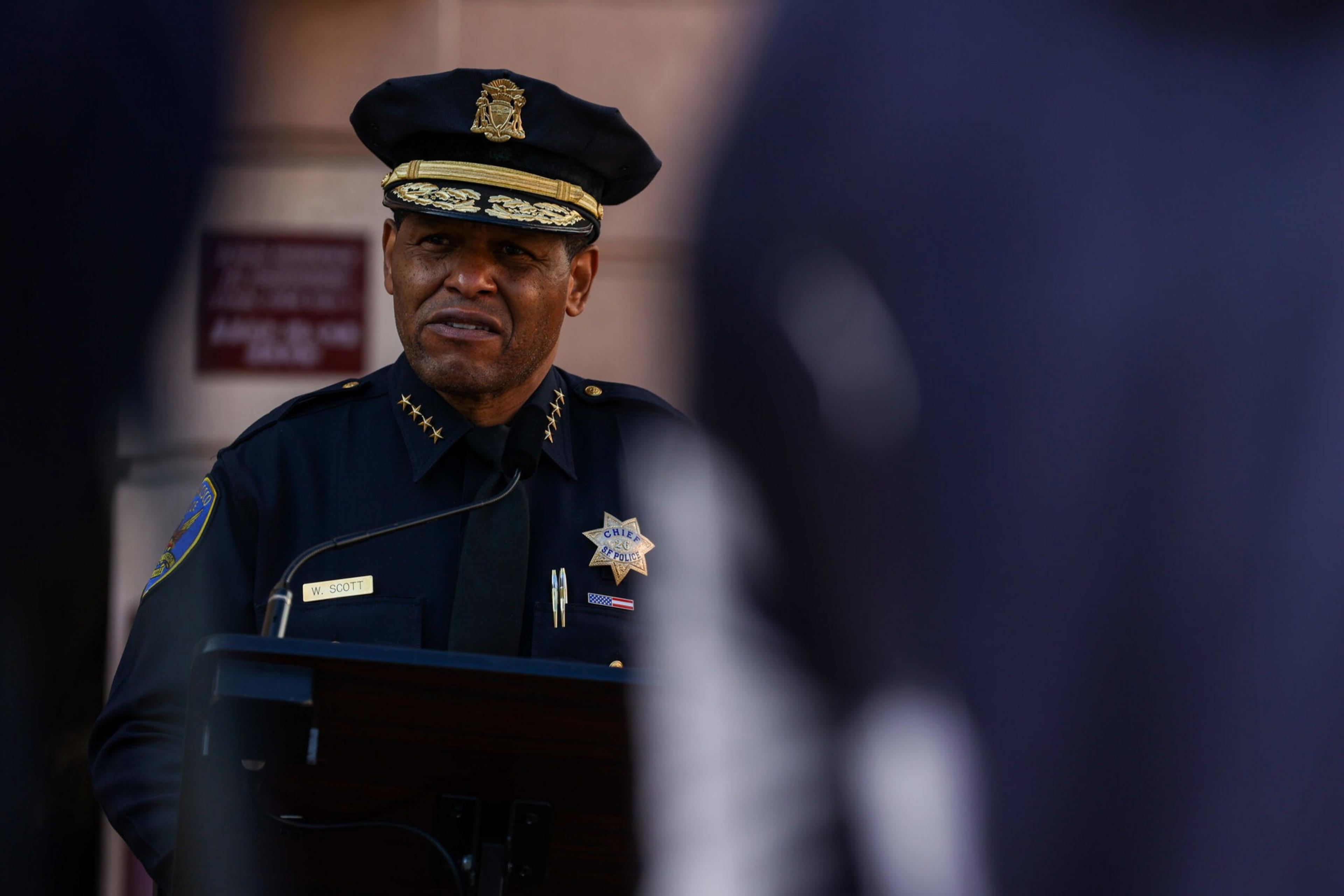 A police chief in uniform with a decorated hat speaks at a podium, framed by blurred figures in the foreground.