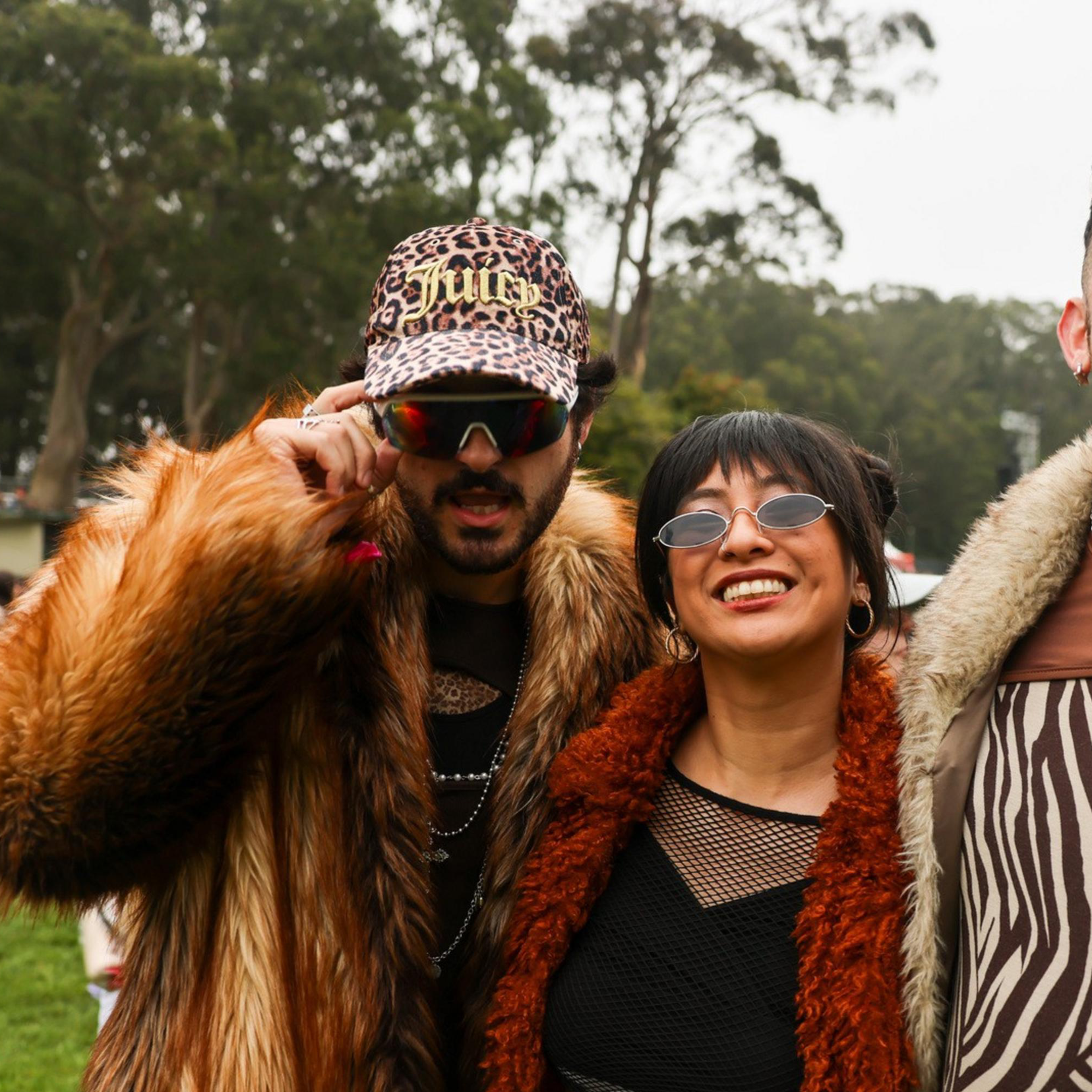 Three people are dressed in colorful, bold outfits, standing happily together outdoors. Trees and a large stage with screens in the background imply a festival setting.