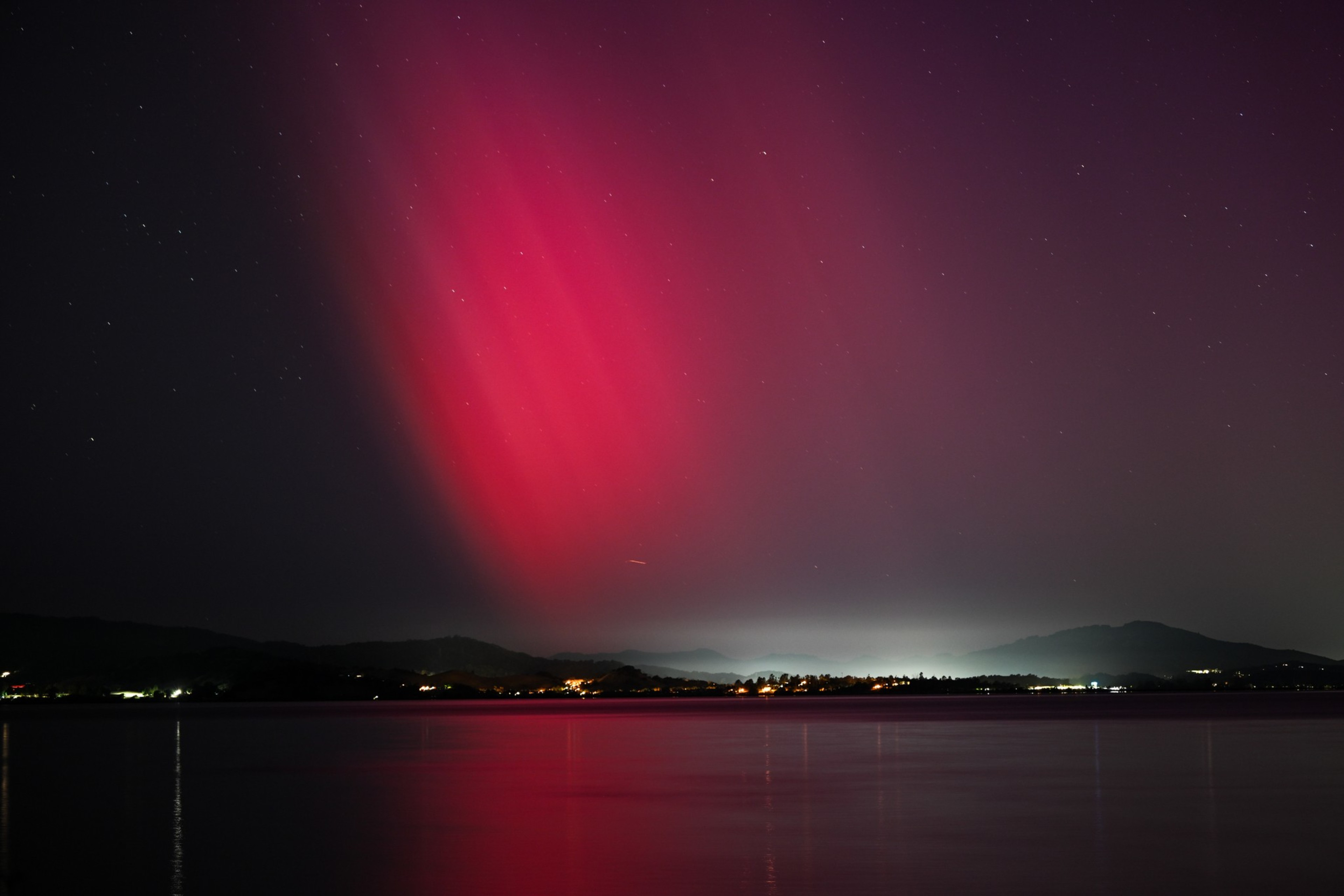 Red aurora-like lights streak across the night sky above glowing city lights and a calm lake.